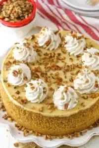 Browned Butter Pecan Cheesecake stunning baked cheesecake with dramatic warm brown butter caramel pecan topping cascading over the edge on a white cake stand on marble
