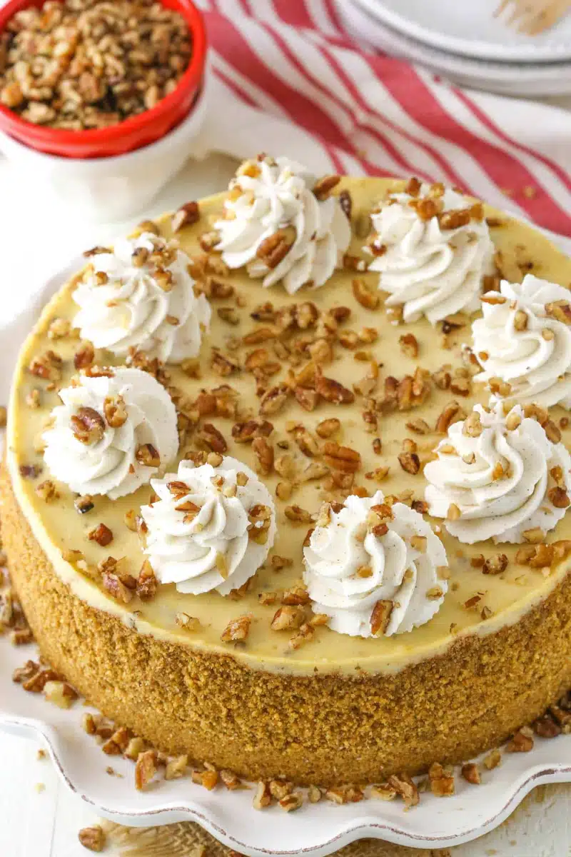 Browned Butter Pecan Cheesecake stunning baked cheesecake with dramatic warm brown butter caramel pecan topping cascading over the edge on a white cake stand on marble